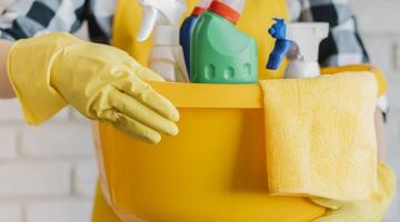 mãos usando luvas seguram um balde com vários produtos de limpeza no interior . #paratodosverem 