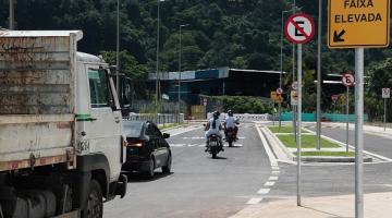 Caminhão passa por via em trecho de curva. Ele passa sobre faixa elevada. Ao lado direito, na calçada a um poste informativo sobre o elevado. Ao fundo se vê a vegetação de um morro. #Paratodosverem