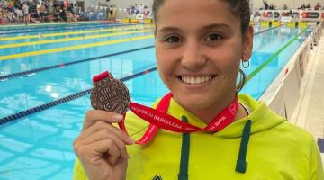 Beatriz Dizotti posa para foto segurando medalha e de costas para uma piscina. #paratodosverem