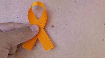 laço laranja está nas pontas dos dedos de uma pessoa ao lado de uma pinta na pele. #paratodosverem