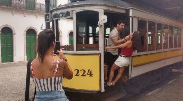 Um casal está nos degraus de entrada do bonde. Um está de frente para o outro. Do lado de fora uma mulher os fotografa com celular. #Pracegover