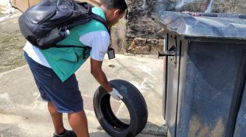 agente uniformizado e usando luvas carrega pneu velho que acumula água. #paratodosverem