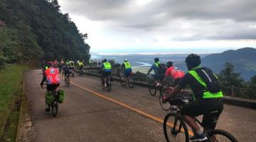 ciclistas pedalam em estrada. Vista da serra dá para a baixada santista. #paratodosverem