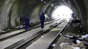 Dois guardas municipais estão dentro de túnel. Há uma pessoa à frente deles carregando um colchão. Nas laterais do túnel há peças de roupas no chão. #Pracegover 