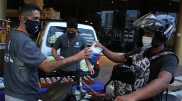 Homem sentado em moto, com capacete e máscara, entrega alimento para homem em pé, também de máscara. #pracegover