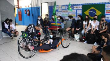 Paratleta mostra cadeira de roda para estudantes e outros observam #paratodosverem