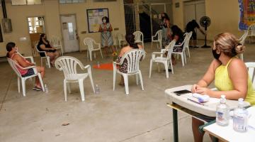 Em primeiro plano uma mulher está sentada tendo junto termômetro e frascos de álcool em gel. Ao fundo, uma roda de conversa. #Paratodosverem