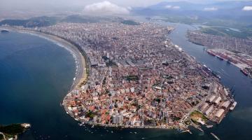 silhueta da ilha de São Vicente, vista do alto, com a ponta da praia em primeiro plano, a orla de Santos do lado esquerdo, e o porto, do direito. #Paratodosverem