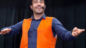 artista está em pé usando um colete cor de laranja e um chapéu coco. Ele está com os braços abertos, falando e usando um microfone preso pela cabeça à frente da boca. #paratodosverem
