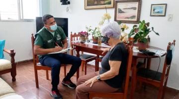 Agente comunitário, uniformizado com colete verde, conversa sentado com senhora na sua sala de jantar. #pracegover