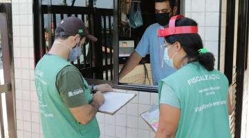 Agentes uniformizados estão em frente a guarita de prédio onde um zelador os atende. #Paratodosverem