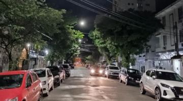 rua iluminada à noite. há carros estacionados. #paratodosverem 