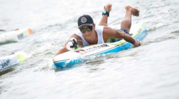 Alex está na água, deitado sobre o paddleboard. #paratodosverem