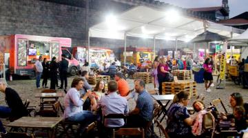 Praça de alimentação com pessoas sentadas e estandes de alimentos. #paratodosverem
