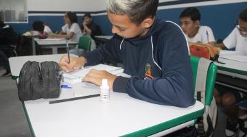 aluno está sentado em carteira escolar escrevendo em caderno. Há outros estudantes ao fundo. #paratodosverem 