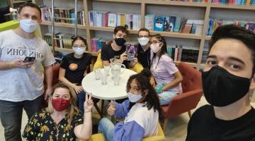 jovens estão sentados em torno de uma mesa. Dois estão em pé. Ao fundo, uma estante cheia de livros. #paratodosverem
