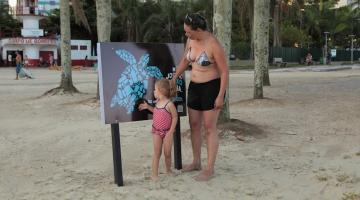 Mulher e criança observam placa educativa instalada na faixa de areia da Praia do José Menino. #pratodosverem