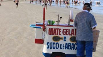 carrinho de mate na faixa de areia com guarda sol aberto. De costas para a foto está o ambulante. #paratodosverem