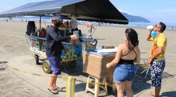 Carrinho de ambulante de bebidas cercado por faixa zebrada. O vendedor atende dois clientes na faixa de areia. #paratodosverem