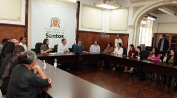 sala de reuniões com várias pessoas sentadas em uma mesa em formato de letra U. Ao fundo, o prefeito ladeado por secretários. Na parede, um banner da Prefeitura com o brasão de Santos. #paratodosverem