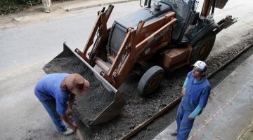 Dois operários trabalham ao lado de escavadeira #pracegover