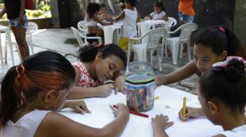 Crianças estão sentadas em torno de mesa. Elas desenham e pintam em folhas de papel. #Pracegover