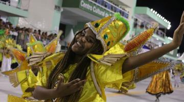 Mulher faz movimento com as mãos para o alto, dançando. Ela tem um adereço na cabeça. #Pracegover