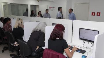 A central de agendamento com pessoas diante de vários nichos de atendimento. Em primeiro plano aparece uma mulher de costas digitando em um teclado de computador. O monitor está à sua frente. #Pracegover