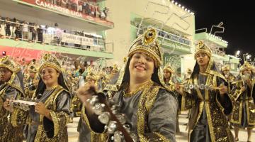 Integrante de bateria movimenta rocar chocalho. Sobre o adereço em sua cabeça se lê: Fé. Ela sorri para a foto Em volta há vários músicos. Ao fundo está a plateia. #Paratodosverem