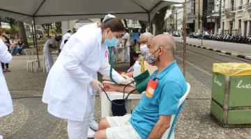 Enfermeira mede pressão de idoso sentado em cadeira na praça #paratodosverem