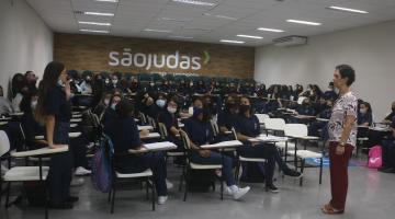 sala cheia de jovens. Uma delas está em pé falando a todas.A coordenadora da roda também está em pé. #paratodosverem