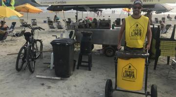 Um carrinho de bebidas está parado na areia da praia. Ele tem um toldo. Em torno dele há  uma lixeira. À frente, um homem segura um carrinho coletor de lixo pequeno. No carrinho e na camiseta do rapaz se lê: Areia Limpa. #Pracegover