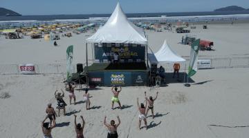 pessoas estão dançando na faixa de areia, diante de uma tenda armada. O mar está ao fundo. #paratodosverem