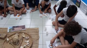 alunos estão sentados no chão desenhando. todos estão diante de cestos com crânios, ossos e outros objetos. #paratodosverem