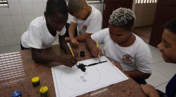 meninos estão pintando cartaz com pincel e tinta. #paratodosverem