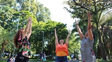 Três mulheres estão em pé em meio a vegetação de parque. Elas estão com os braços para cima, como num movimento de ioga, de inspiração. Olham para o alto. #Pracegover