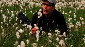 jornalista está no meio de um campo de flores com uma câmera fotográfica na mão. Ao fundo, um morro. #paratodosverem