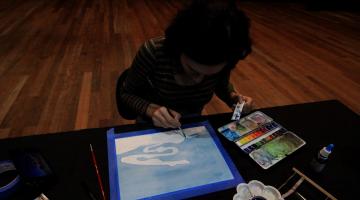 mulher está pintando aquarela sobre uma tela de papel em uma mesa. Ao redor dela há uma paleta de titantes e vários petrechos de pintura. #paratodosverem