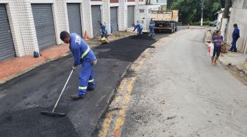 trecho de rua é asfaltado. Há homens ajustando o asfalto e a máquina compactadora ao fundo. #paratodosverem