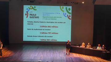 duas pessoas em pé em um palco. Uma projeção ao fundo com texto e quatro pessoas sentadas a uma mesa também no palco. #paratodosverem 