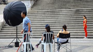 equipamentospara filmagem e fotografia dispostos em praça. Ao fundo, numa escadaria há uma mulher parada, de perfil, indicando que é a modelo alvo da filmagem. #paratodosverem