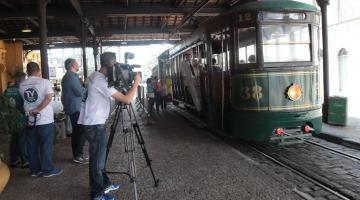 Equipe está na estação de bonde pegando imagens do bonde que está parado. Um dos homens manuseia a câmera que está montada em tripé. #paratodosverem