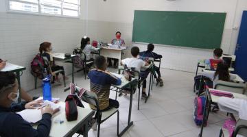 sala de aula com professor ao fundo e alunos sentados às carteiras. Lousa e ventilador afixados na parede ao fundo. #paratodosverem