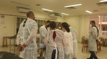 pessoas vestindo avental branco estão em pé em torno de uma mesa em uma sala. Não é possível ver o que há sobre a mesa. #paratodosverem