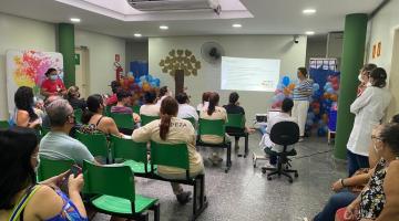 público está sentado em sala assistindo palestra. Ao fundo, uma imagem é projetada na parede. A sala está decorada com balões coloridos. #paratodosverem