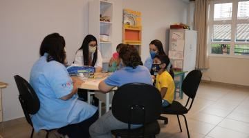 várias pessoas sentadas em torno de uma mesa. Há crianças, jovens e adultos. Ambiente é de uma escola. #paratodosverem