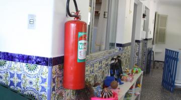 extintor de incêndio afixado em parede de escola. Abaixo, um móvel aberto, com vários recipientes com brinquedos. #paratodosverem