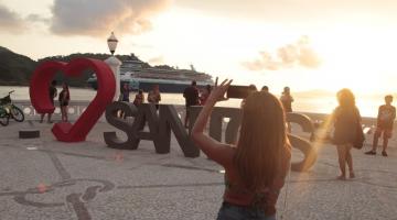 mulher etá diante de monumento Santos fotografando. Há um navio passando ao fundo. #paratodosverem