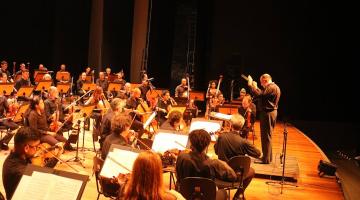 os músicos da orquestra no palco e o regente à frente. #paratodosverem 