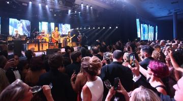 pessoas dançam em salão diante de palco onde músicos se apresentam.Há holofotes sobre eles. #paratodosverem 